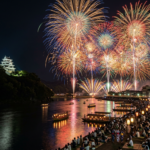 Fireworks lighting up the night sky over a river with a Japanese castle and crowds watching from the riverbank