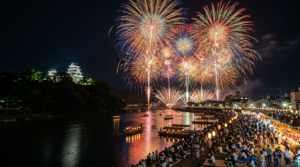 Fireworks lighting up the night sky over a river with a Japanese castle and crowds watching from the riverbank